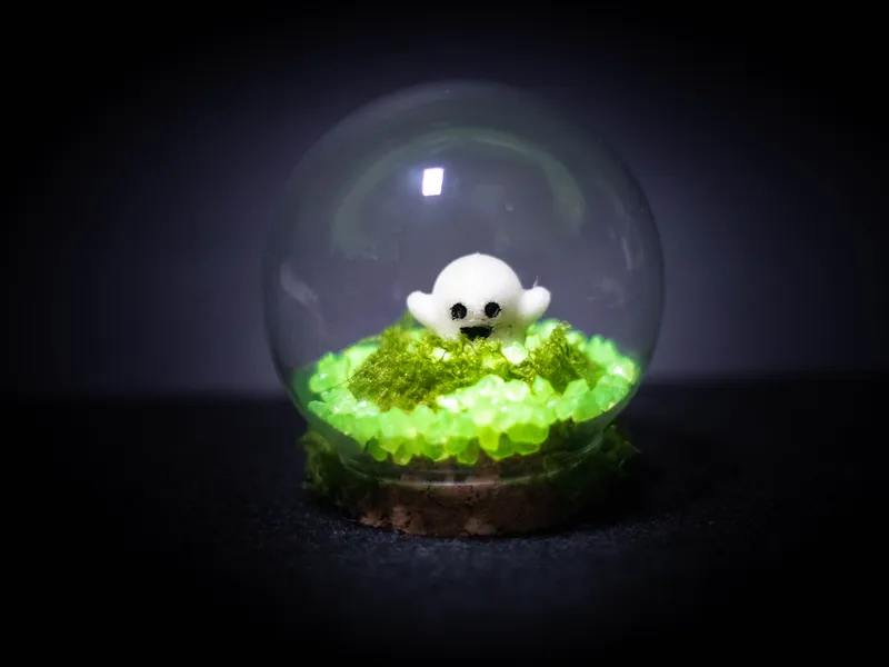 Glass dome terrarium with a small white ghost on moss and glowing green stones